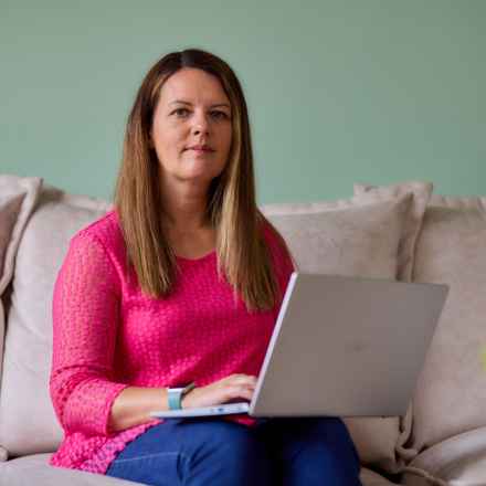 Claire wearing a pink top and blue jeans sits in her living room, using her laptop