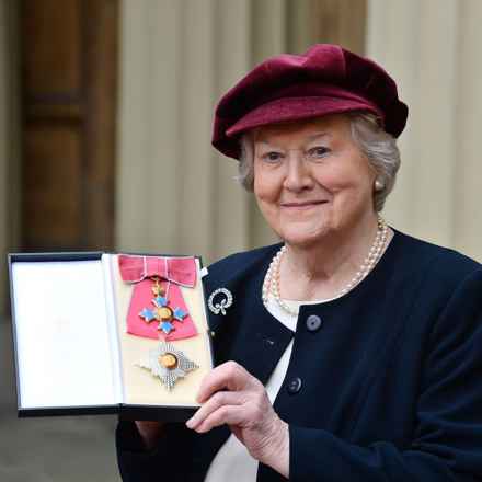 Patricia, in a hat and smart clothes, smiling holding her OBE