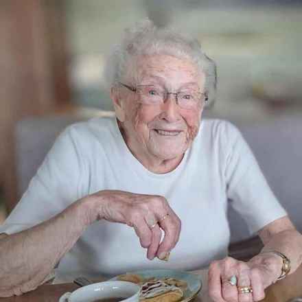 Joan, who passed away in 2024, wearing a white top pictured smiling while enjoying her cup of tea and baked treat