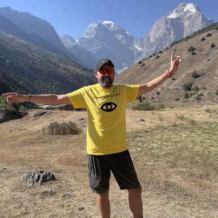 Richard wearing his yellow Macular Society t-shirt holds his hands in the air, with mountain scenery in the distance.