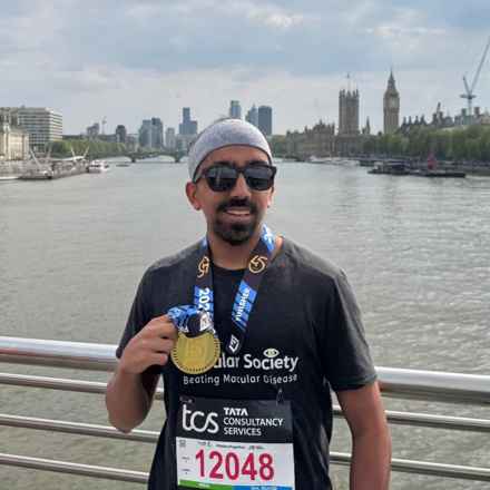 2025 London Marathon runner Armaan stands on a bridge spanning the River Thames, smiling as he holds his runner's medal. The London skyline, including Big Ben, can be seen in the background.