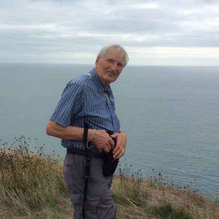 John smiling as he walks along the cliffs, with view across the sea behind him.