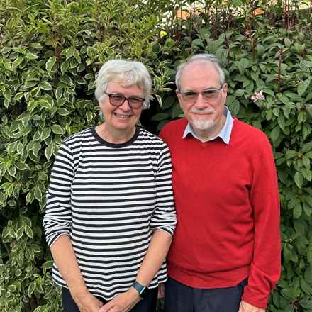 Jane, left, wearing glasses and a horizontal striped jumper, standing next to her husband Ed. He is also wearing glasses and a red jumper.