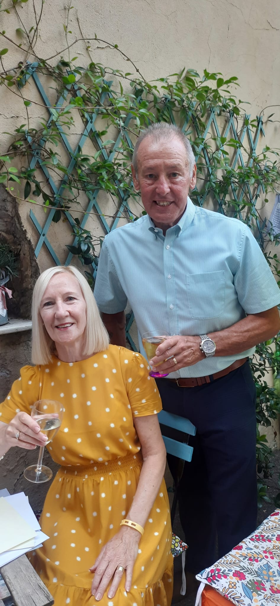 Peter and wife sue posing to camera. Peter standing smiling wearing a light blue shirt, with his arms around wife sue who is sat down wearing yellow spotted dress